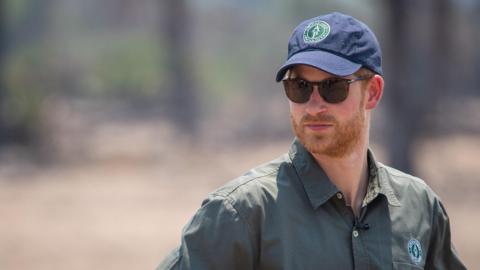 Prince Harry staring to his right wearing an African Parks cap and shirt with sunglasses