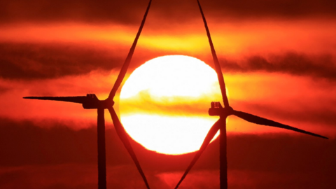 The image shows two wind turbines silhouetted against a vivid sunset. The sun is positioned directly between the blades of one of the turbines, creating deep orange and red tones in the sky.