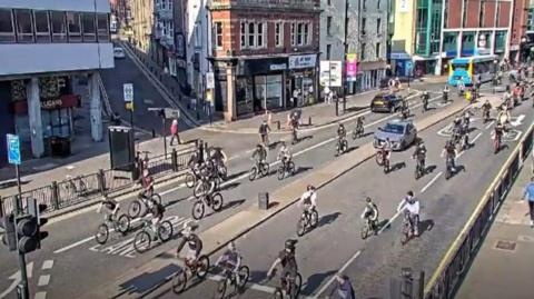CCTV footage showing more than 20 people on bikes on both sides of a road, which has a central reservation. There is a bus travelling down the road on the left. There are buildings towards the back of the photos with various shopfronts on the ground floor.