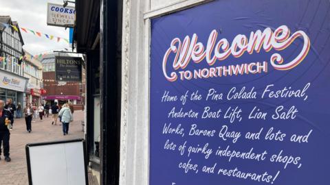 A large purple-and-white sign in a high street doorway which reads: "Welcome to Northwich, Home of the Pina Colada Festival, Anderton Boat Lift, Lion Salt Works, Barons Quay and lots of lots of quirky independent shops, cafes, and restaurants!"