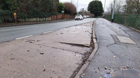 A photo of a road, concrete layby and footpath, looking downhill towards a bend. There are large cracks in the layby and smaller cracks on the footpath. To the left of the road, a weathered brick wall stands in front of thick green foliage. To the right, green railings stand above an embankment, with bare trees rising behind it. Cars can be seen in the middle distance.