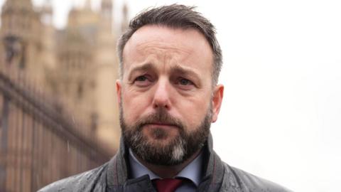 Colum Eastwood. He has short dark hair and beard, wearing a dark raincoat, white shirt and red tie.