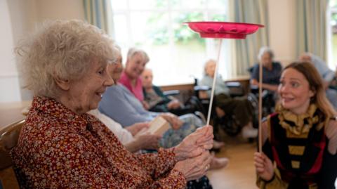 An elderly woman, sitting in a care home, holds a spinning plate on a stick. In the background, other older people are watching her.