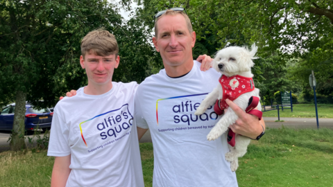 A young man is smiling while standing next to a man who is also smiling and holding a small white dog. Both the teenager and the man are wearing Alfie's Squad tops.