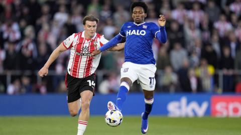 Ipswich Town's Jaden Philogene, wearing blue shirt, white shorts and blue socks, controls the ball with his left foot while being pursued by Southampton's James Bree, wearing a red and white striped shirt, black shorts and white socks.