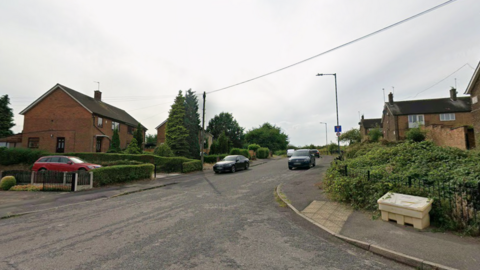 A Google streetview of a cul-de-sac with cars parked on both sides and houses.
