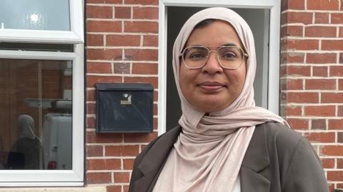 A woman wearing glasses and a headscarf stands outside a house smiling for the camera.