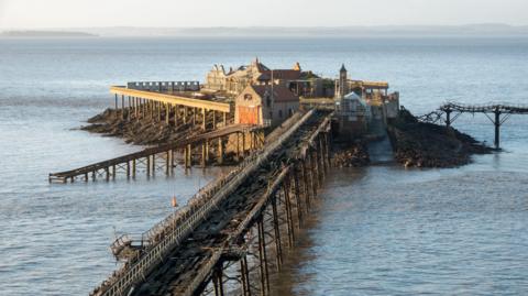 Birnbeck Pier in Weston-super-Mare. The pier is damaged and in disrepair, with a heavily damaged wooden walkway and dilapidated buildings.