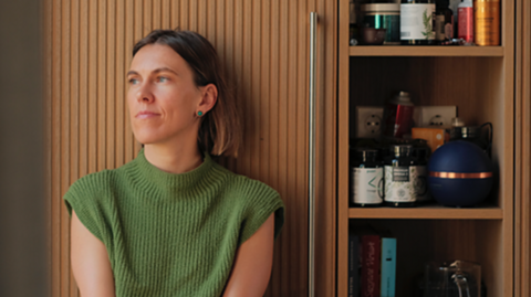 A woman with short brown hair, dressed in a textured green knit top, she stands or sits beside a tall wooden cabinet. 
The cabinet’s vertical grooves and partially open door reveal a curated interior: shelves lined with supplement containers, jars, books.