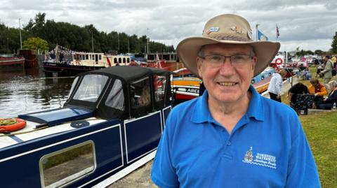 David wears an Australian type hat with a blue polo shirt on with Yorkshire Waterways logo on it. Canal boats can be seen in the background on the water.