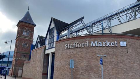 A curved brick wall on a building with a clock tower behind it. There is a glass ceiling and tiled atrium.