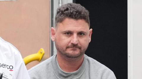 A head-and-shoulders shot of a man with a stubble beard and short, brown, wavy hair. He is wearing a grey jumper.