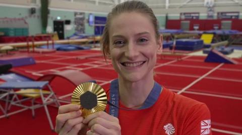 A smiling Bryony Page posing with her Paris 2024 Olympic gold medal. She has fair hair which is tied back. She is wearing a red Team GB top and is standing in a large sports hall with a red floor. Behind her are a number of blue trampolines and ramps.