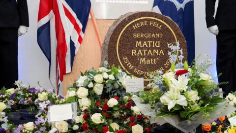 A stone saying 'Here Fell Sergeant Matiu Ratana "Matt", surrounded by flowers and hand-written cards