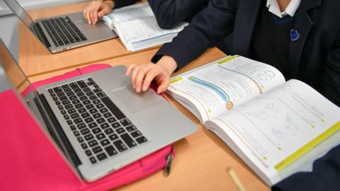 Two children sit side by side. They have text books and laptops in front of them and appear to be doing school work.