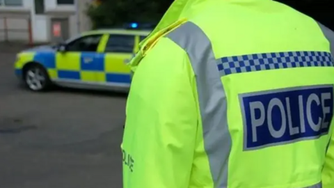 A Greater Manchester Police officer wearing a hi-vis jacket. They have their back to the camera. A blurred out police vehicle is in the background.