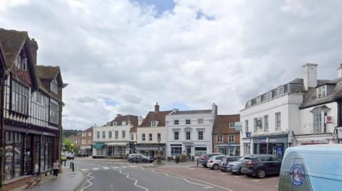 A google Streetview image showing Westerham High Street with shops either side and a zebra crossing across the road. There are cars parked in spaces on the right of the image and most buildings are two and three storeys high.