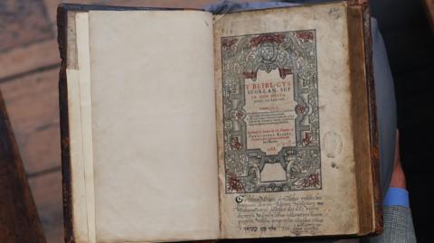 A man in a grey suit with grey hair holds an antique-looking book. The photo is captured from above so the viewer looks down on the scene. The man is stood on a wooden floor with a red carpet laid down. The book has a beautiful Celtic design on the right hand page and calligraphy. The paper in the book is yellowed and spotted with age.