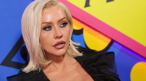 Christina Aguilera at The Abbey 35th Anniversary Party held at The Abbey on May 20, 2026 in Los Angeles, California. She is wearing a black top and has shoulder length blonde hair. Christina is standing in front of a brightly coloured blue, yellow and pink backdrop.