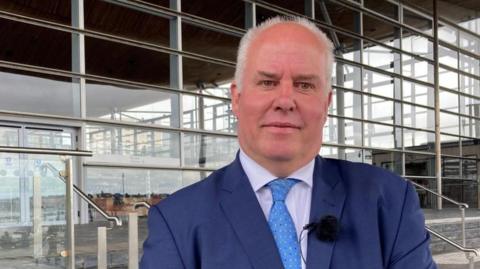 Andrew RT Davies stood in front of the Senedd wearing a dark blue suit and a light blue tie.