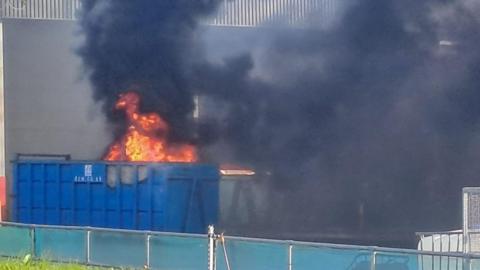 A fire is seen burning inside a large blue metal container on an industrial site, with a lot of black smoke billowing from it.