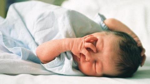 Image of a newborn baby asleep in a hospital maternity ward. The baby has dark hair on its head.