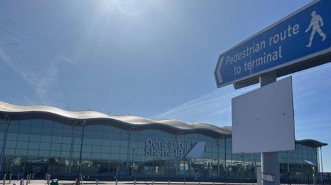 Blue pedestrian route sign pointing towards Doncaster Sheffield Airport terminal building, a glass fronted building with a wavy roof. Bright sun and blue sky is above.