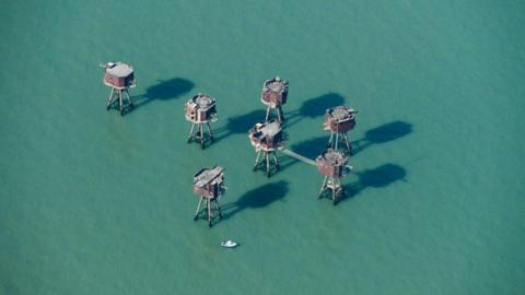 An aerial view showing the Redsands forts in the middle of the sea. There are seven of them and they are octagonal shaped structures rising out of the water on long metal legs. They are brown and rusting and a small boat is in the water near to them.