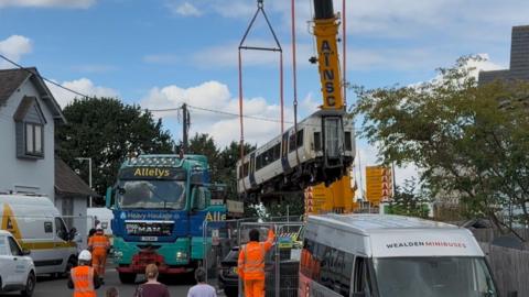 A crane carrying one of the derailed carriages as a small group of members of the public and workers in hi vis jackets and hard hats watch. A few vans and vehicles are parked to the sides and there are trees and housing nearby.