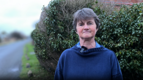 Lesley Parker is standing in front of a bush outside a house by the road. She is wearing a blue jumper and has short, grey hair.