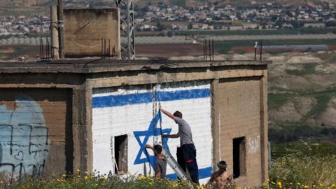 Israeli right-wing activists paint an Israeli flag on the side of an abandoned building in the Jordan Valley, in the occupied West Bank, with neighbouring Jordan visible in the background (11 March 2024