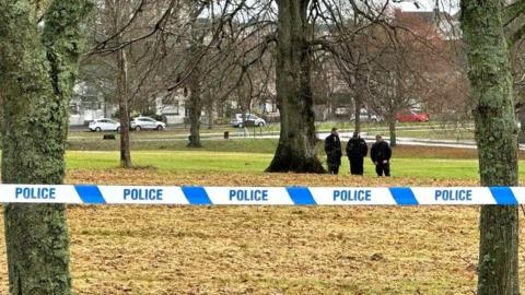Blue and white police tape between trees in a park, with police officers in the background.