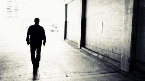 A black and white backlit silhouette of a man walking through an urban setting. The wall next to him is made of bricks.