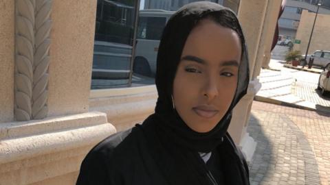 Marjama Osman is seen in a black hijab and abaya standing outdoors, looking directly at the camera in bright sunlight.