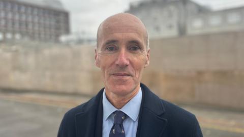 Deputy Renouf is a man with a bald head, staring into the camera. He is wearing a navy coat, with a blue shirt and navy tie. He is standing in front of a wall with buildings out-of-focus behind him.