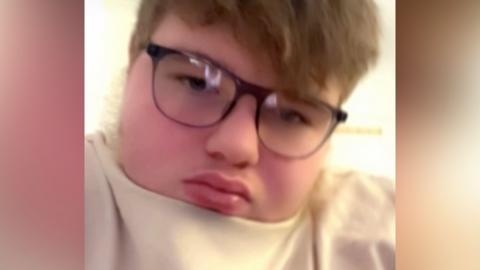 A boy with mousey hair wears a cream top and dark-rimmed spectacles while looking at the camera.