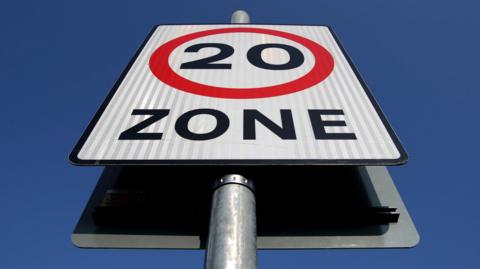 A photo of a 20mph zone sign, taken from below, with a clear blue sky behind it. 