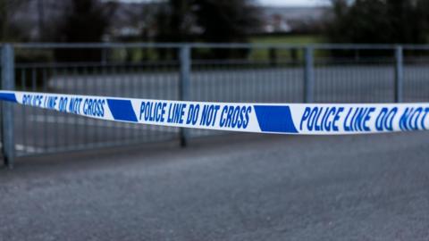Blue and white police tape with a grey fence in the background.
