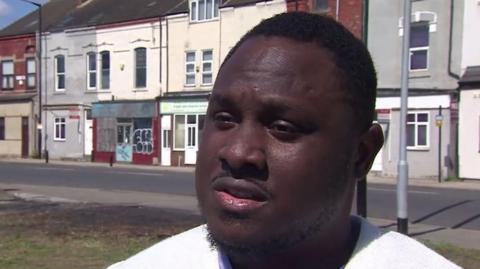 Carer Brendan Nwabichie being interviewed with the scorched grass in the background where his car was set on fire