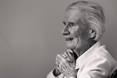 A black and white photo of an elderly lady sitting in a chair against a plain wall, smiling with her hands clasped as she looks out of the frame.