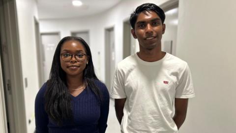 Two young people, one male with black hair and a white T-shirt, and one female with long black hair, glasses and a dark blue jumper are stand in a corridor smiling at the camera