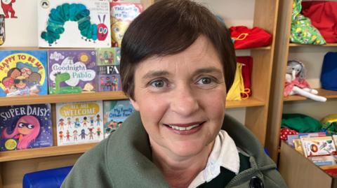 Tracey Roleston smiling at the camera. She has short brown hair and is wearing a green top. Behind her are bookshelves with books and toys.