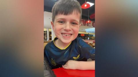 Archie Benwell, a seven-year-old boy with brown hair, smiles at the camera. He is wearing a Newcastle football shirt.