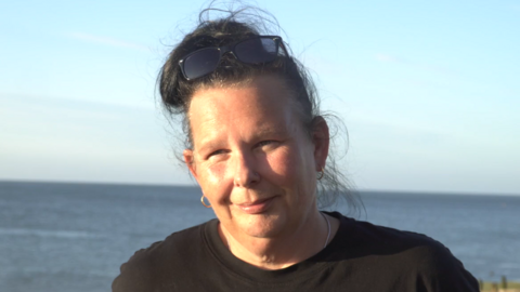 A woman standing with the beach and sea in the background. She has her long, black hair tied up in a bun, is wearing a black t-shirt and has black sunglasses on her head.