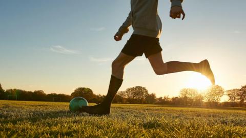 A young person is about to kick a football on a grass field. In the background is a line of trees, and the sun is glaring above them.