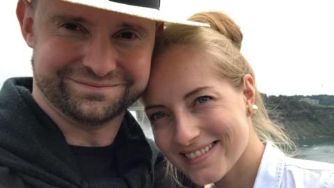 Christopher Jones is on the left and is wearing a black T-shirt, black jumper and a white hat. He has a short brown beard and moustache. Standing next to him on the right is his wife Paula, who has blonde hair in an updo and is wearing a white shirt.