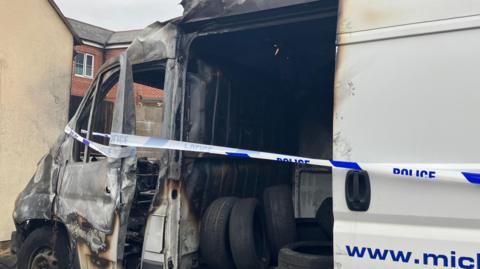 A burned out white van full of tyres. The inside of the van has been blackened by fire damage. Police tape is covering the open doorway and is wrapped around the van's window, which no longer has any glass in it. The front end of the van is now a grey and black colour due to the damage.