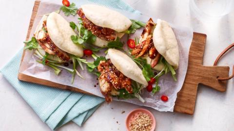 Four bao buns sit on baking paper on a wooden chopping board. The white buns are filled with marinated chicken, sliced chillis and coriander