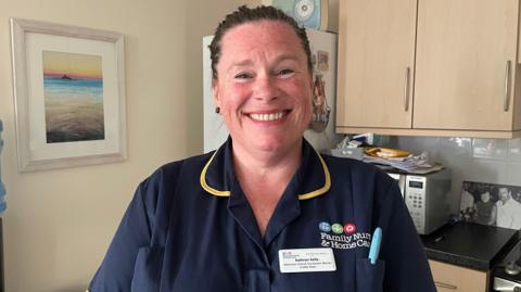 A nurse called Kathryn Kelly smiles broadly while standing in a kitchen. She is wearing a dark blue uniform with Family Nursing & Home Care's logo on it. She also has a white name badge on.