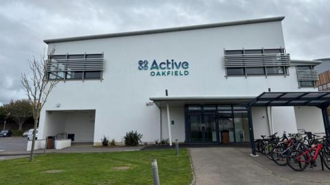 Bicycles are parked in a rack outside the entrance to a two-storey white building. it has the words Active Oakfield on it in blue letters. There is a path leading to the double door and a small patch of grass to the left of the image.